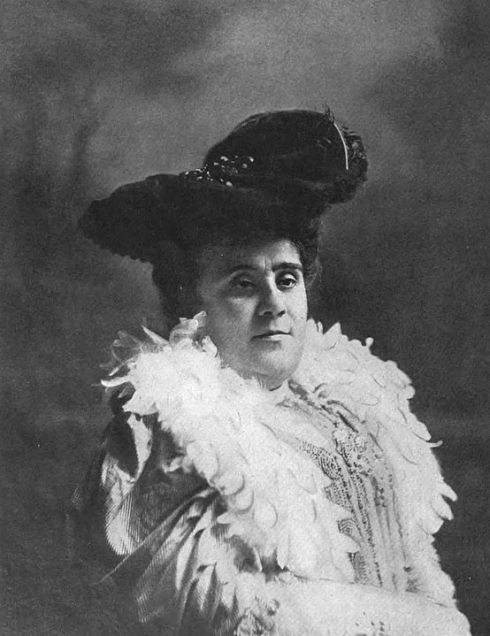 A photo of Matilda Serao in mid-life (perhaps in her 40s or 50s). She is dressed affluentially. She is wearing a hat and a feather shawl and her dress is elaborate and costly. She is in a fixed pose of the era. 