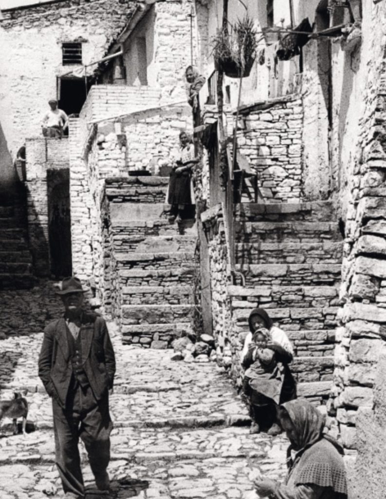 A Lacedonia street scape - stone built walls and stairs. A man stands near a sitting woman holding a child. Frank Cancian image.