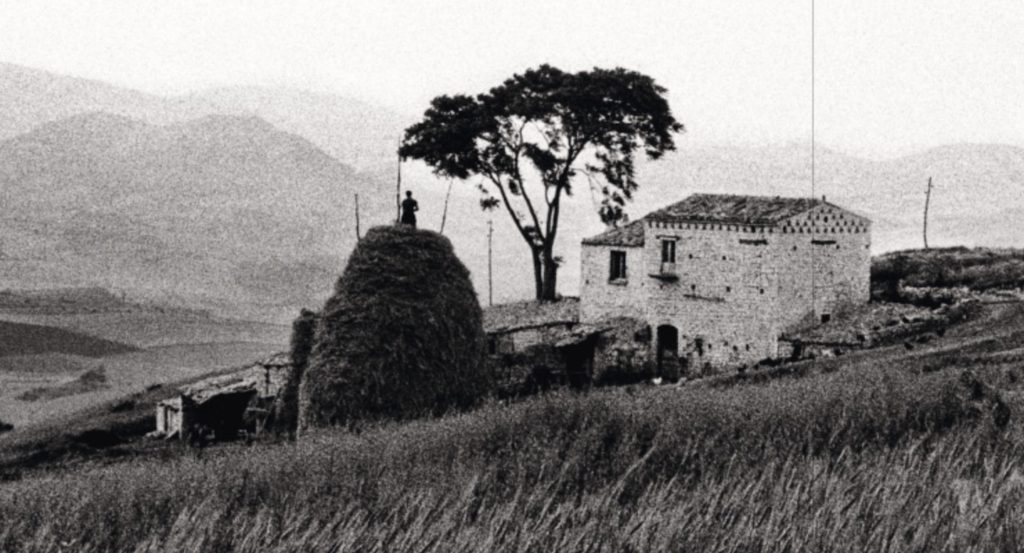 A view of the country side near Lacedonia. A man stands in the distance on a large conical haystack, almost taller than the building which is to its right. Frank Cancian image.